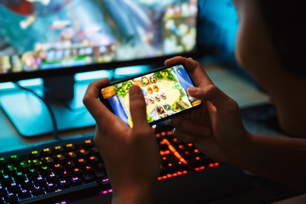 Retrato de muchacho adolescente jugador jugando videojuegos en el teléfono inteligente y la computadora en una habitación oscura, usando audífonos y usando un teclado colorido retroiluminado