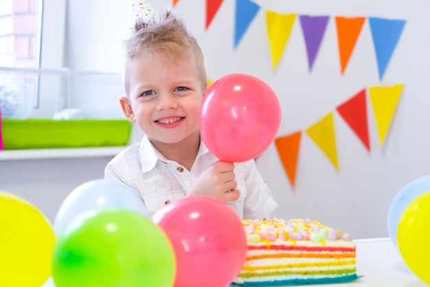 Retrato del muchacho caucásico rubio que sonríe en la cámara cerca de la torta de cumpleaños del arco iris. Fondo colorido festivo con globos