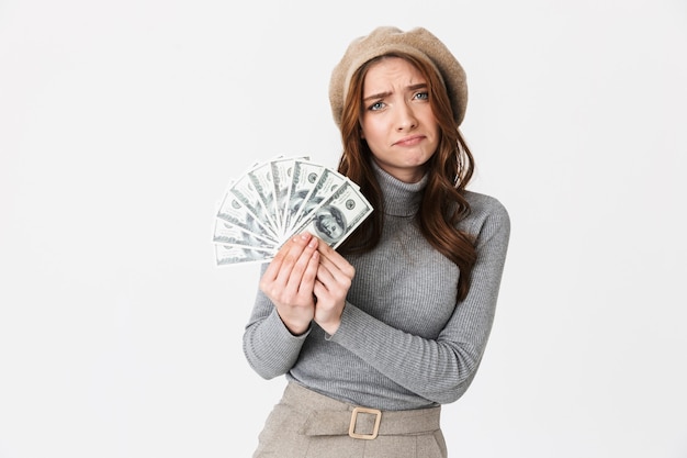 Retrato de mujer de 30 años con el ceño fruncido con sombrero sosteniendo ventilador de dinero en dólares aislado en blanco