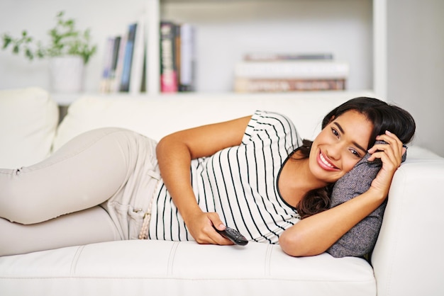 Retrato de una mujer acostada en el sofá con el control remoto de la televisión y viendo la televisión con una sonrisa en la cara en la sala de estar Es hora de relajarse transmitiendo películas hogar brillante mujer feliz en el sofá con televisión en el salón en India