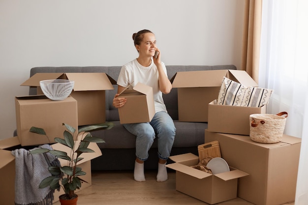 Retrato de una mujer adorable con peinado de moño con pantalones blanco casual y jeans, sentada en un sofá, rodeada de cajas durante la mudanza, hablando por teléfono móvil.