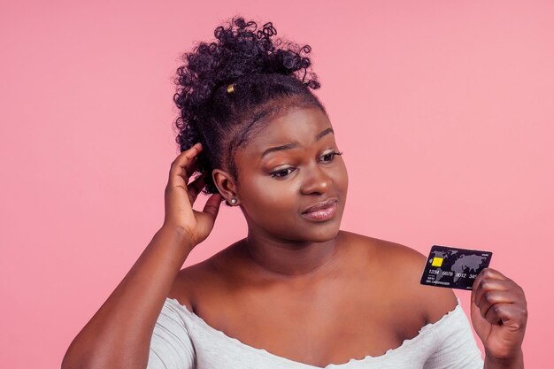 Retrato de una mujer adorable con el pelo peludo sosteniendo una tarjeta de crédito de plástico y sonriendo sinceramente aislada sobre el fondo del estudio rosa.