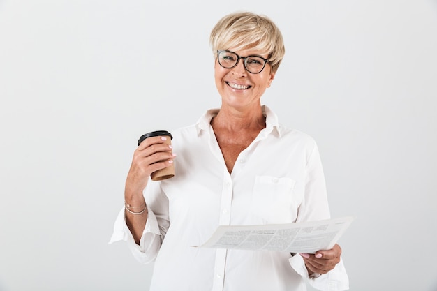 Retrato de mujer adulta alegre con anteojos sosteniendo una taza de café para llevar y leyendo el periódico aislado sobre la pared blanca