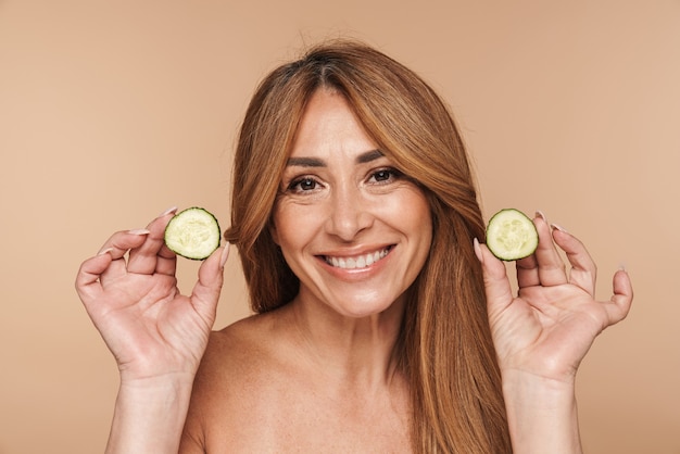 Retrato de mujer adulta sin camisa sosteniendo pepino y sonriendo aislado en beige