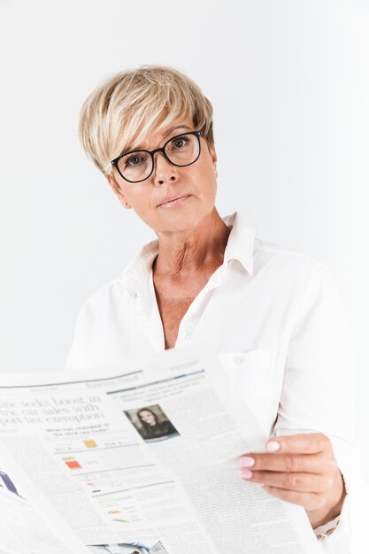 Retrato de mujer adulta enfocada usando anteojos leyendo el periódico aislado sobre una pared blanca en el estudio