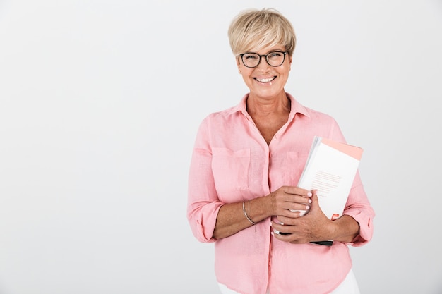 Retrato de mujer adulta feliz con anteojos sosteniendo libro de estudio aislado sobre una pared blanca en el estudio