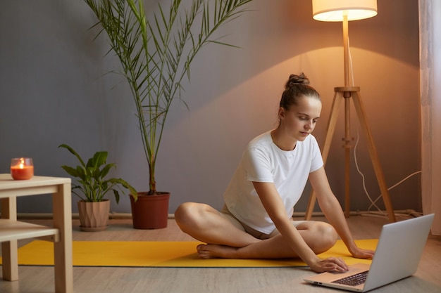 Retrato de mujer adulta joven concentrada con moño de pelo con camiseta blanca practicando yoga usando una computadora portátil e internet para elegir un curso en línea