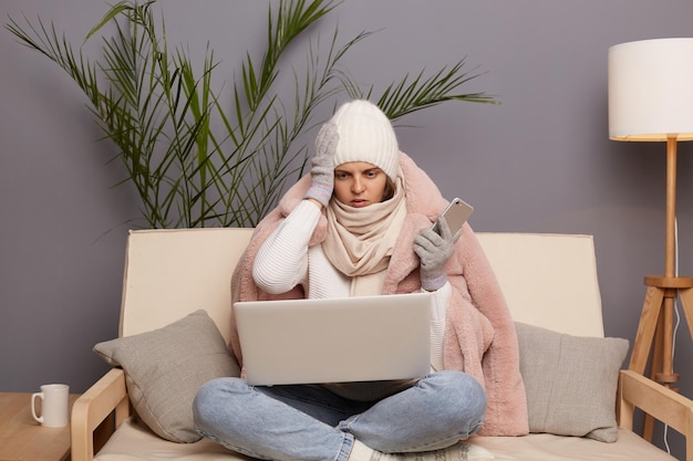 Retrato de una mujer adulta joven conmocionada posando en una habitación fría sentada en la tos con guantes calientes y gorra trabajando en línea en una laptop que tiene problemas con su trabajo