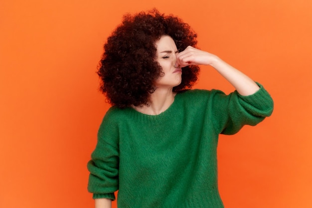 Retrato de una mujer adulta joven con peinado afro que usa un suéter de estilo casual verde que contiene la respiración con los dedos en la nariz olor apestoso pedo Estudio interior aislado en fondo naranja