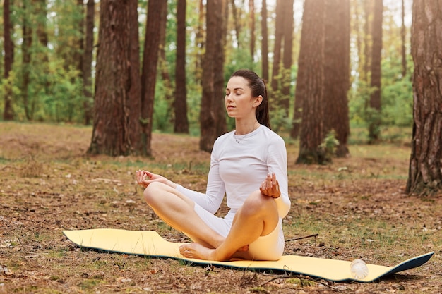 Retrato de mujer adulta joven en top blanco y leggins sentada en la estera con las piernas cruzadas en posición de loto y meditar, manteniendo los ojos cerrados