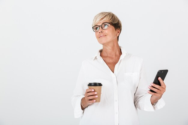 Retrato de mujer adulta pensativa con anteojos sosteniendo teléfono móvil y taza de café para llevar aislado sobre una pared blanca en el estudio