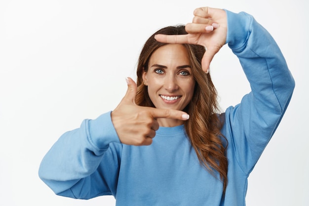 Foto retrato de una mujer adulta sonriente y segura de sí misma de 30 años que mira a través de los marcos de la mano feliz mirando algo tomando fotos de pie contra el fondo blanco