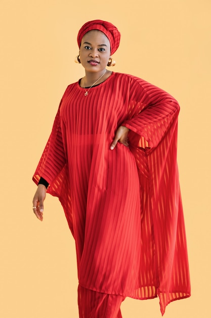 Retrato de mujer africana segura con elegante vestido rojo y turbante en la cabeza, sonriendo a la cámara con el brazo en la cintura, posando sobre fondo amarillo.