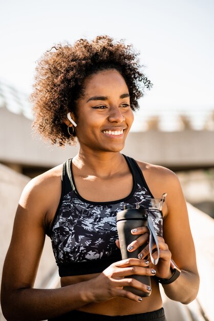 Retrato de mujer afro atleta sosteniendo una botella de agua y relajarse después de hacer ejercicio al aire libre