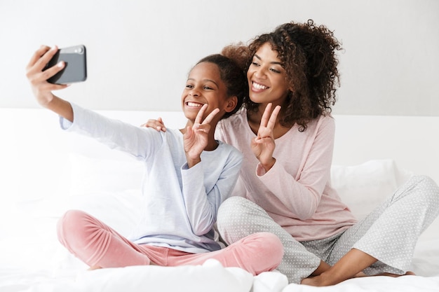 Retrato de mujer afroamericana alegre y su pequeña hija teniendo retrato selfie en teléfono móvil en casa