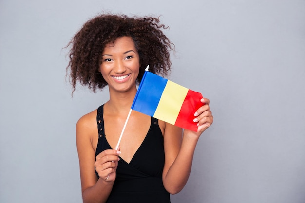 Retrato de una mujer afroamericana feliz sosteniendo la bandera rumana sobre la pared gris y mirando al frente