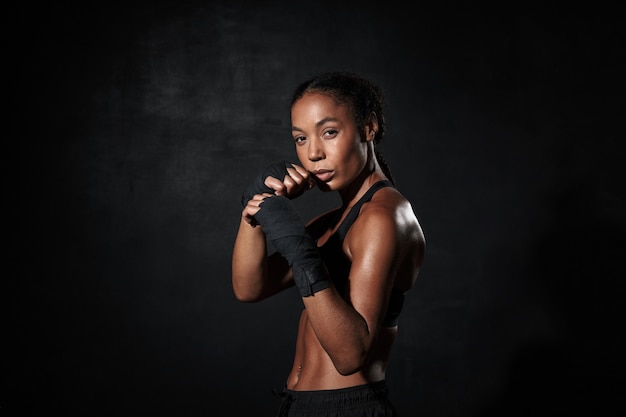 Retrato de mujer afroamericana fuerte con ropa deportiva de boxeo en la mano envuelve aislado en negro
