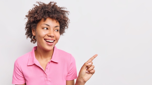 Retrato de mujer afroamericana de pelo rizado sonriente apunta el dedo índice a la derecha muestra banner de venta de descuento viste camiseta rosa casual aislada sobre pared blanca