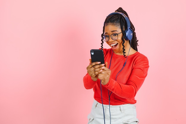 Retrato de una mujer afroamericana relajada con auriculares escuchando música y usando un teléfono móvil aislado sobre fondo rosa