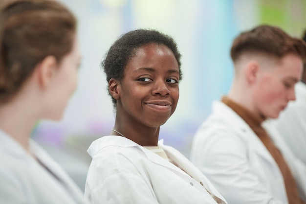 Retrato de mujer afroamericana sonriente con bata de laboratorio mientras está sentado en la fila en la audiencia y escucha una conferencia sobre medicina en la universidad, espacio de copia