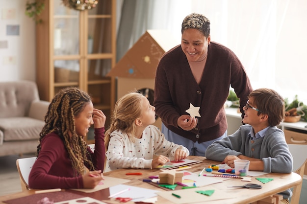 Retrato de mujer afroamericana sonriente enseñando clase de arte con un grupo multiétnico de niños haciendo tarjetas de Navidad hechas a mano en la escuela