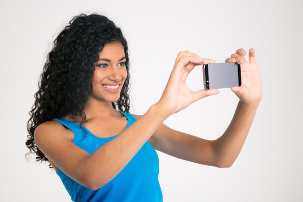 Retrato de una mujer afroamericana sonriente haciendo foto selfie en smartphone aislado en una pared blanca