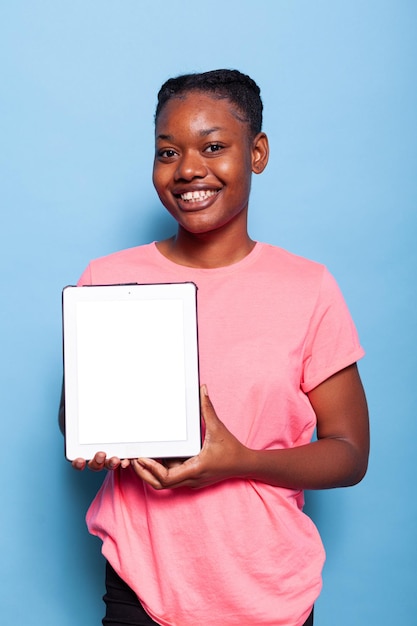 Retrato de una mujer afroamericana sonriente sosteniendo una tableta con pantalla blanca de pie en un estudio con fondo azul. Adolescente que recomienda la aplicación ipad de publicidad. Panel táctil aislado