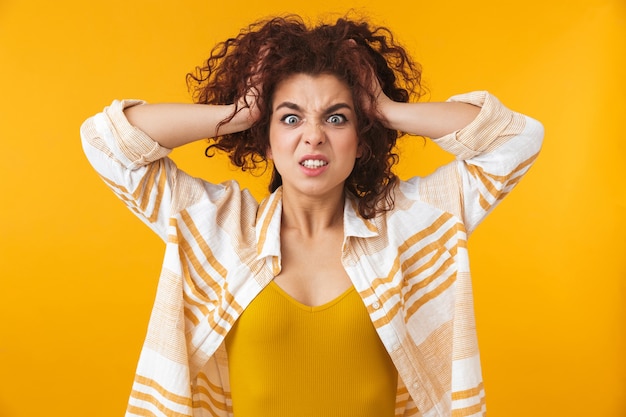 Retrato de mujer agresiva de 20 años con cabello rizado agarrando su cabeza, aislado en amarillo