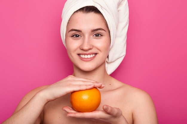 El retrato de la mujer alegre atractiva con la toalla blanca en la cabeza, sostiene la naranja sobre la pared rosada. Joven sonriente visita el salón de spa y descansa, cuida su piel. Concepto de belleza natural.