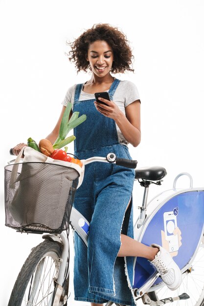 Retrato de mujer alegre celebración de teléfono celular mientras monta en bicicleta con bolsa de compras aislado sobre la pared blanca