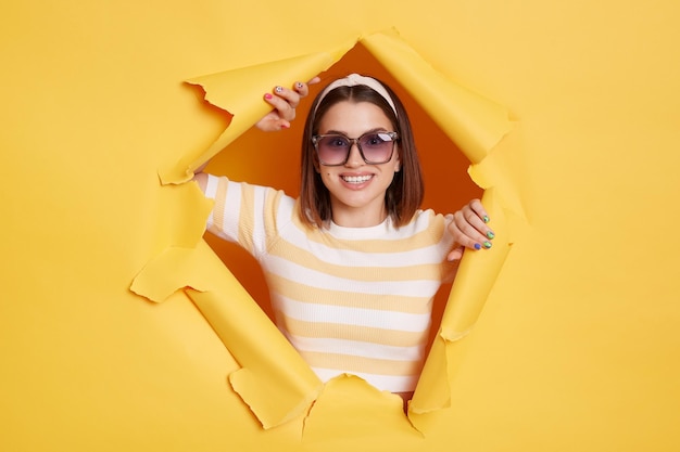 Retrato de una mujer alegre y encantadora que lleva una banda de pelo de camiseta a rayas y gafas de sol se rompe a través de un fondo de papel amarillo en un agujero rasgado que expresa felicidad mirando a la cámara