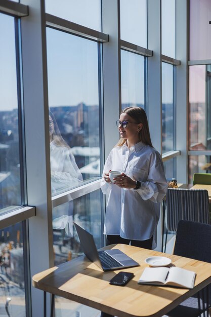 Retrato de una mujer alegre con gafas clásicas sonriendo en su tiempo libre en un café con café mujer judía positiva en un escritorio de camisa blanca con trabajo remoto portátil