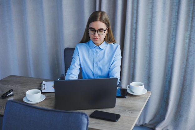 Retrato de mujer alegre en gafas clásicas sonriendo en tiempo libre en café con café mujer europea positiva en escritorio de camisa azul con trabajo remoto portátil