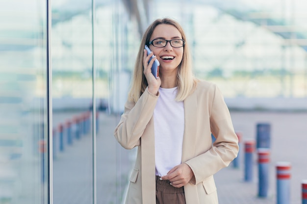 Retrato de una mujer alegre con gafas rubia sosteniendo un teléfono móvil sonriendo hablando por un teléfono inteligente cerca de la oficina
