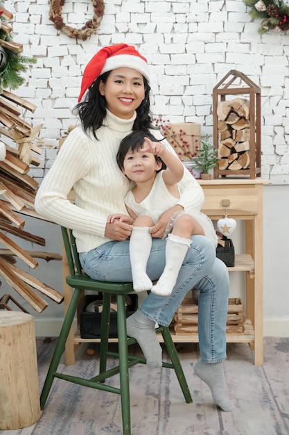 Retrato de mujer alegre con gorro de Papá Noel sentado en un taburete con hija pequeña en regazos