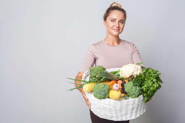 Retrato de mujer alegre con moño con canasta de verduras frescas y mirando a cámara, dieta vegetariana, comida sana, espacio de copia para publicidad. tiro del estudio de interior, fondo gris