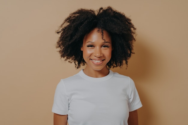 Retrato de mujer alegre de piel oscura que expresa felicidad mientras está de pie sobre fondo beige