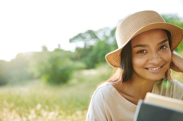 Retrato de mujer alegre con piercing de labios y sombrero de paja libro de lectura mientras está sentado sobre el césped en el parque verde