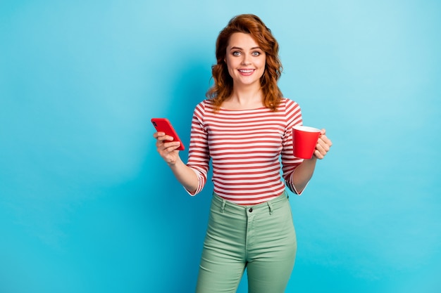 Retrato de mujer alegre positiva sostenga la taza con café con leche, use mensajes de texto de teléfono inteligente escribiendo notificación de red social use pantalones verdes pantalones aislados sobre color azul