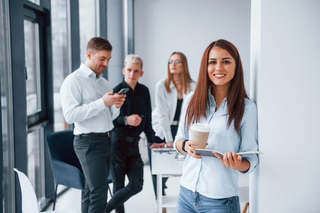Retrato de una mujer alegre con una taza de bebida que se para frente a un joven equipo exitoso que trabaja y se comunica juntos en el interior de la oficina
