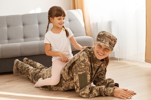 Retrato de mujer alegre con uniforme de camuflaje y gorra posando con su hija soldado mujer tendida en el suelo con su pequeño niño feliz en la espalda