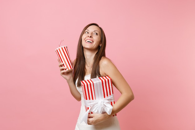 Retrato de mujer alegre en vestido blanco mirando hacia arriba mantenga caja roja con regalo, presente, taza plactic con cola o soda