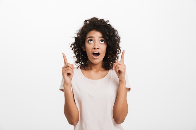 Retrato de mujer americana en camiseta básica con cabello rizado mirando hacia arriba y señalando con el dedo índice en copyspace, aislado sobre la pared blanca