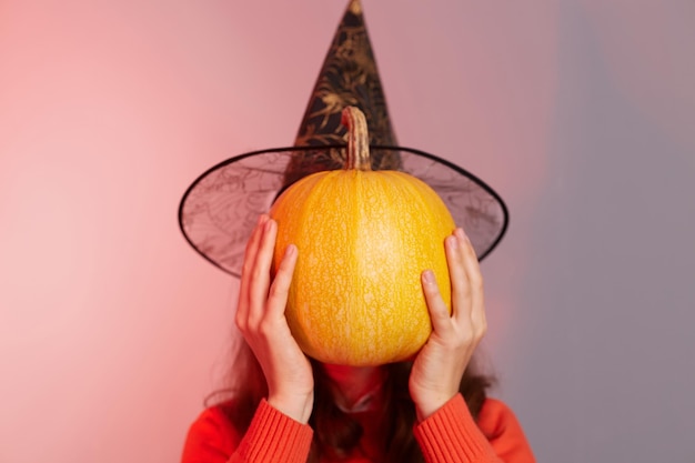Retrato de una mujer anónima desconocida escondiendo su rostro detrás de una gran calabaza naranja madura que se cubre con suéter y sombrero de bruja aislado sobre un colorido fondo de luz de neón