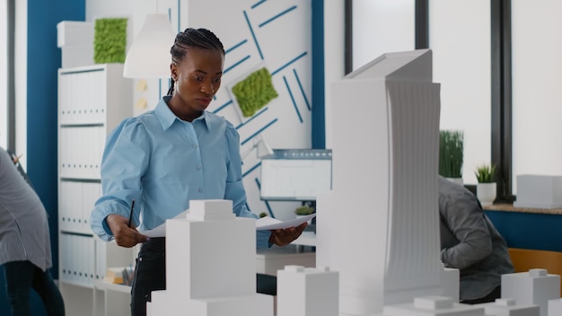 Retrato de mujer arquitecta mirando el plan de planos para encontrar inspiración para el diseño de la construcción. Ingeniero trabajando con modelo de construcción y maqueta para diseñar proyecto de arquitectura urbana.