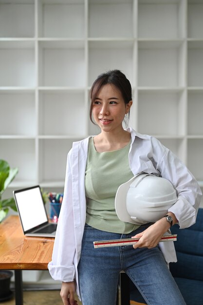 Foto retrato de mujer arquitecto en camisa de pie en la oficina y sosteniendo el casco en sus manos.