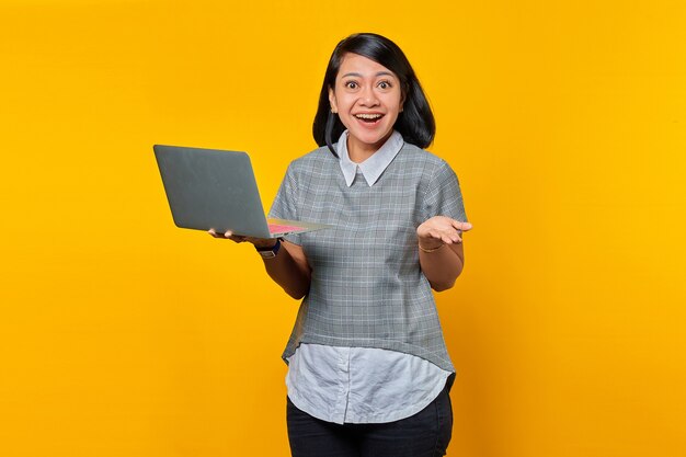 Retrato de mujer asiática alegre con gafas sosteniendo portátil y levantando la mano sobre fondo amarillo