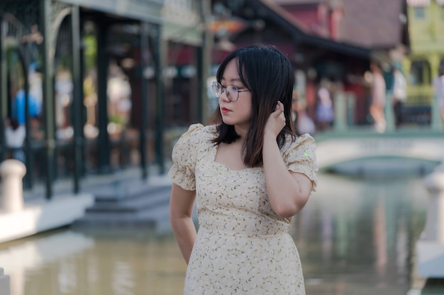 Retrato de mujer asiática con anteojos tocar su cabello y pensar cerca del río en la colorida ciudad, mujer en ropa casual relajante, vergonzosa mujer mirando a otro lado