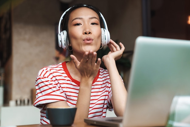 Retrato de mujer asiática atractiva con auriculares soplando beso de aire mientras usa la computadora portátil en el café en el interior