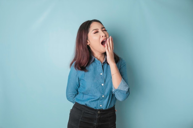 Retrato de una mujer asiática atractiva y soñolienta que usa una camisa azul y se siente cansada después de la noche sin dormir bostezando